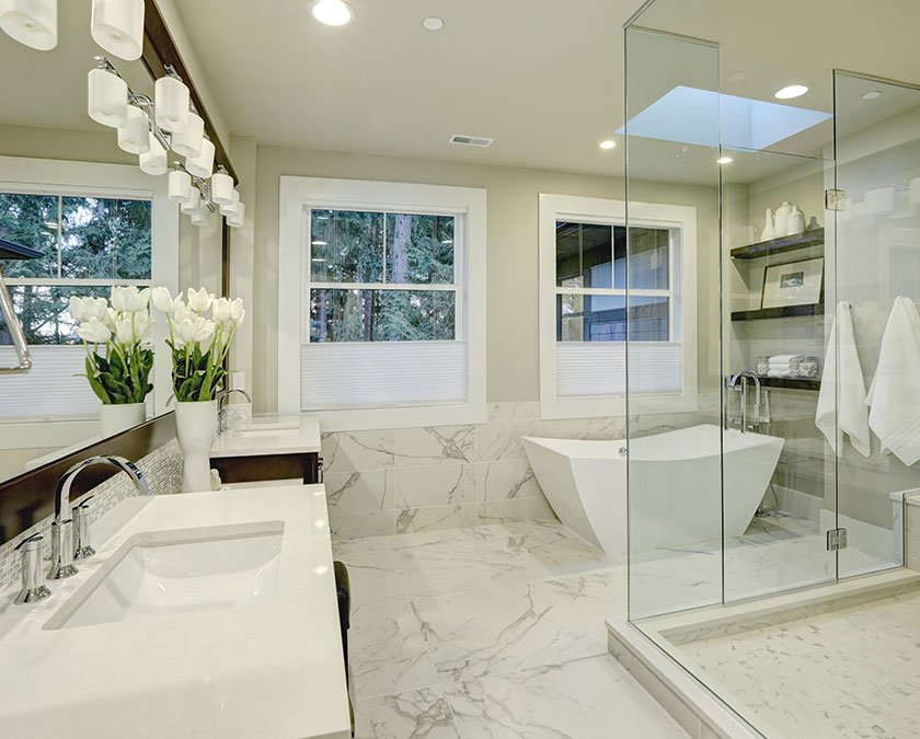 Modern bathroom with while marble on the floor and walls, modern bathtub, wooden cabinets with white countertops, large mirror, LED lights, and a glass tower shower.
