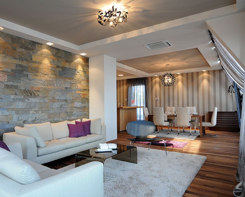 Modern living room with stone decor on the main wall, wooden floor, white carpet, white couch, and open space dining area with white chairs.
